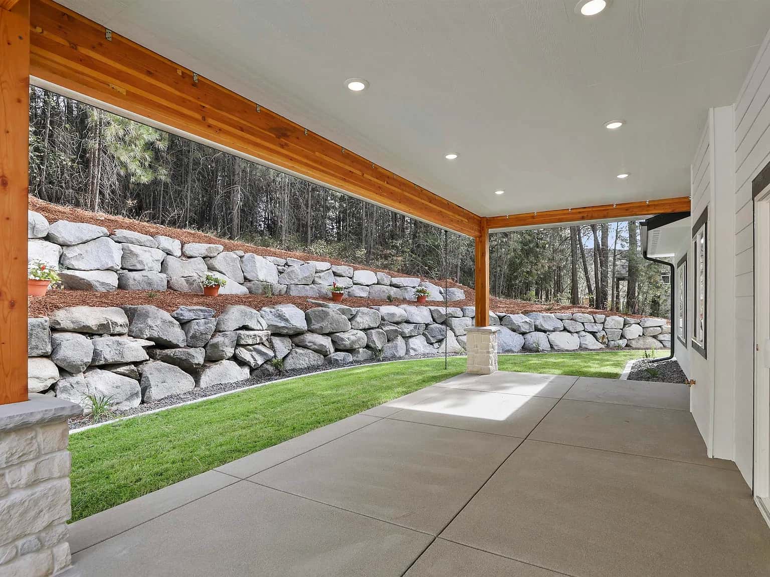 Covered back patio with timber beams looking onto a boulder retaining wall and pines
